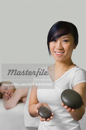 Nahaufnahme eines Massage-Therapeuten halten zwei Kieselsteine mit einer reifen Frau hinter ihr auf einem Massagetisch liegend