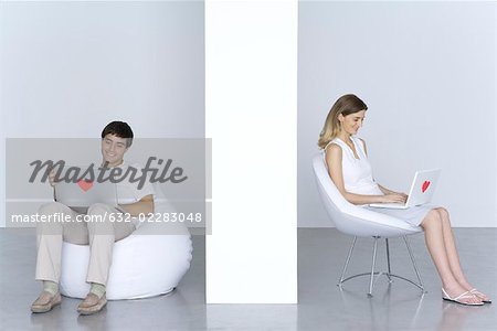 Man and woman sitting separately, using laptop computers with hearts on them, smiling