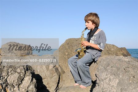 Boy Playing Saxophone on Rocks by Shoreline