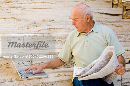 Man Reading Newspaper and Using Laptop Computer