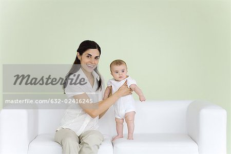 Woman helping baby stand on couch, smiling at camera