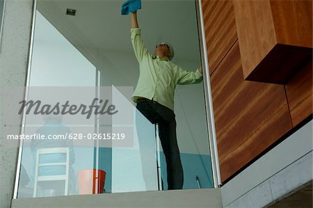 Senior man cleaning a window pane next to a senior woman who is sitting on a chair, low angle view