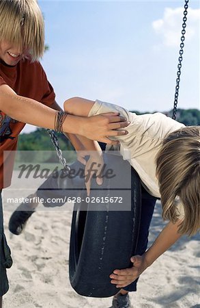 Blonde Boy pushing a Rubber Tire-Swing on which his Friend lies procumbently - Friendship - Fun - Playground