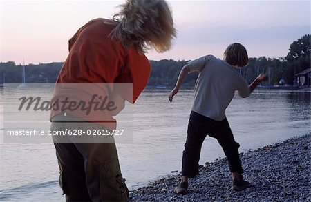 Two Boys throwing flat Stones over the Water Surface - Smartness - Game - Friendship - Lake - Dusk