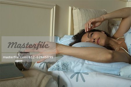 Young woman lying in bed, silencing alarm clock