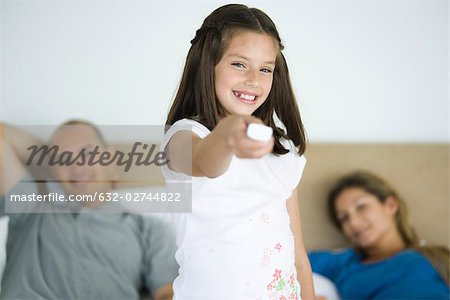Girl holding out remote control, smiling at camera, parents in background