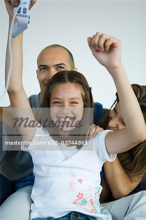 Girl sitting with parents, holding video game controller in air and smiling