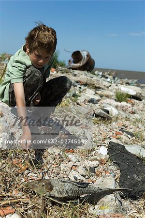 Boy crouching on polluted shore, poking dead fish with stick