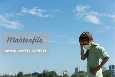 Boy standing outdoors wearing pollution mask, holding head, city in background