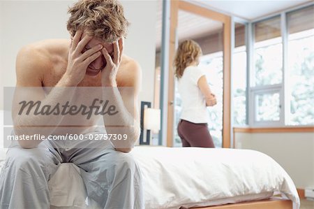 Man Sitting on Edge of Bed and Woman looking out Window in Background