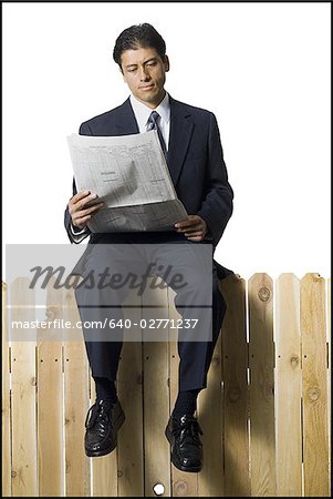 Businessman sitting on fence reading newspaper