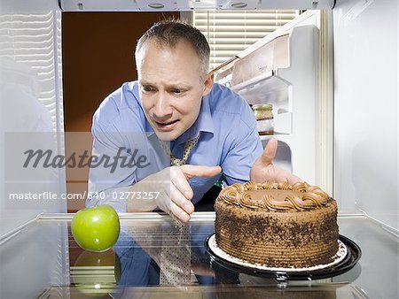 Man in refrigerator reaching for green apple looking at chocolate cake