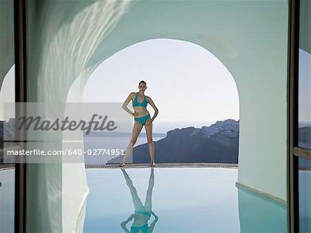 Woman in bikini standing at edge of infinity pool with arch and rock formation