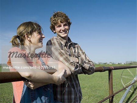 Brother and sister leaning on wooden fence in field with blue skies