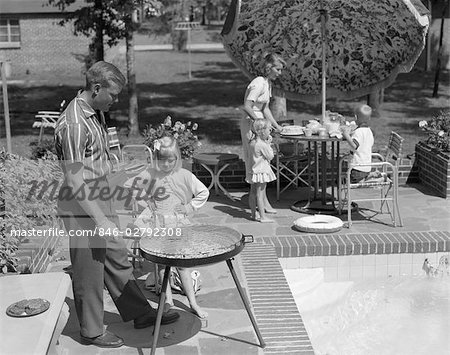 1960s FAMILY OF 5 ON THE PATIO FATHER IS GRILLING AS DAUGHTER WATCHES MOTHER SET UMBRELLA COVERED TABLE