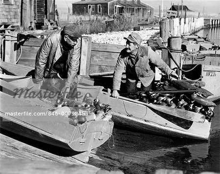 1930s 1940s TWO MEN DRESSED FOR DUCK SHOOTING PUSHING OFF FROM SHORE IN TWO SMALL BOATS LOADED WITH DUCK DECOYS SHOTGUN