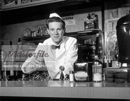 ANNÉES 1950 SOURIANT JEUNE HOMME SODA JERK SE PENCHE AU COMPTOIR OFFRANT UNE GLACE AU CÔNE LOOKING AT CAMERA