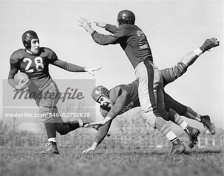 ANNÉES 1940 TROIS HOMMES JEU DE FOOTBALL EN CUIR CASQUES UN HOMME S'ATTAQUE À UN AUTRE QUI ESSAIE D'INTERCEPTER LA TROISIÈME EN PLEIN AIR