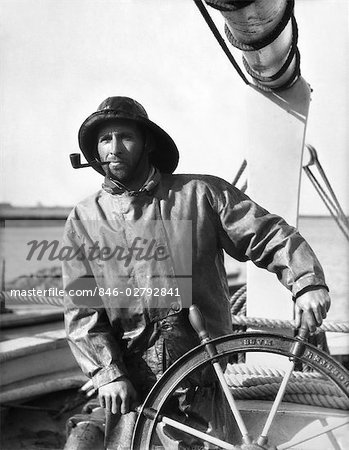 1930s MAN SAILOR FISHERMAN AT THE HELM OF FISHING BOAT WEARING FOUL WEATHER GEAR AND SMOKING A PIPE