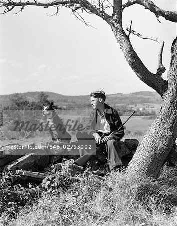 1940ER JAHRE BOY AUF STEIN ZAUN MIT HUND RUHEN GEWEHR ZWISCHEN BEINEN BOSTON TERRIER SITZEND