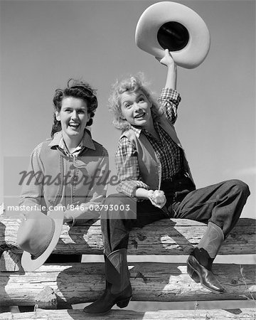 1940ER JAHRE ZWEI LÄCHELNDE MÄDCHEN IN COWBOY-OUTFITS AUF EINEM ZAUN SITZEND IST IN LUFT AUSLÖSEN HAT ANDERE STEHEN NEBEN IHR