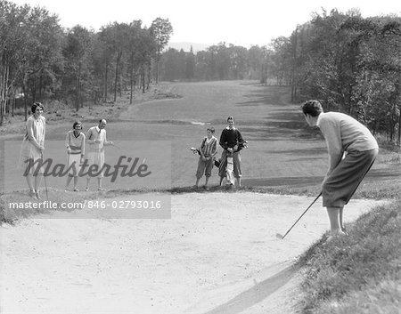 1930s MAN IN KNICKERS GOLFING IN SAND TRAP WITH MEN & WOMEN LOOKING ON