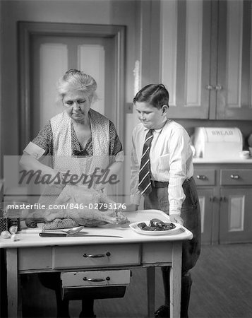 1930s GRANDMOTHER IN APRON PREPARING A TURKEY IN THE KITCHEN WHILE GRANDSON IN DRESS SHIRT TIE & KNICKERS WATCHES