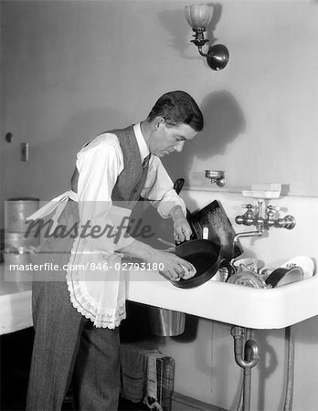 1930s MAN DRESSED IN SHIRT TIE VEST & APRON BENT OVER A SINK FULL OF DISHES CLEANING AN IRON SKILLET WITH A DISHCLOTH