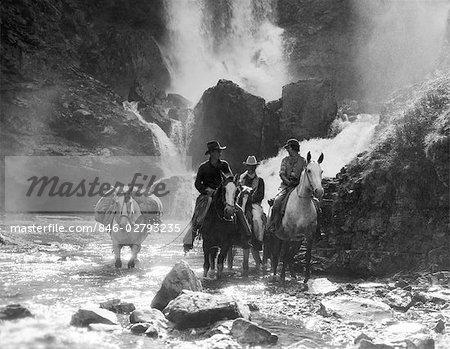 ANNÉES 1930 DEUX HOMMES ET FEMME ÉQUITATION À CHEVAL AVEC LE QUATRIÈME CHEVAL TRANSPORTANT DES FOURNITURES EN FLUX