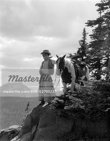 1940s MAN COWBOY IN CHAPS VEST HAT AND KERCHIEF WITH PALOMINO HORSE ON PEAK OVERLOOKING GLACIER PARK MONTANA USA
