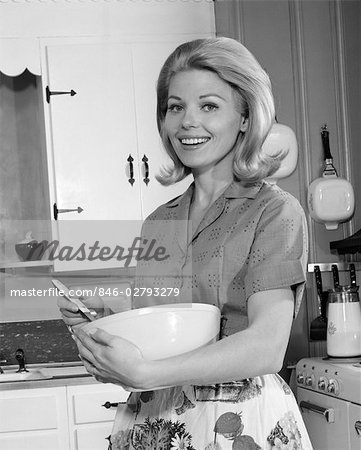 1960s 1970s SMILING BLOND WOMAN HOUSEWIFE IN KITCHEN HOLDING MIXING BOWL AND SPOON