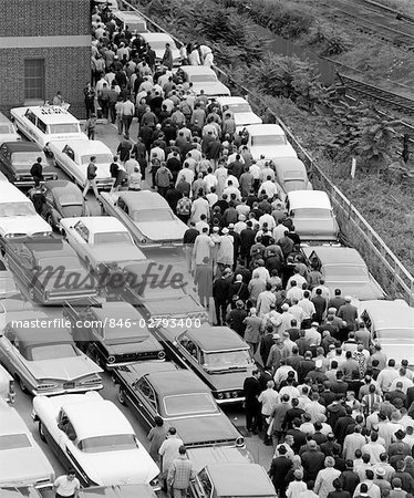 1960s OVERHEAD OF FACTORY WORKERS IN CROWDED PARKING LOT GETTING OUT OF CARS & WALKING TOWARD BUILDING TO WORK