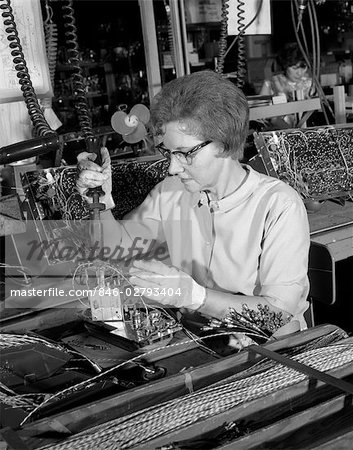 GROS PLAN DES ANNÉES 1960 DE FEMME PORTANT DES LUNETTES & DE TRAVAIL GANTS À SOUDER DES PIÈCES POUR LE MONTAGE DE TÉLÉVISEURS COULEUR