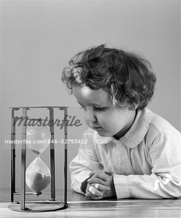 1940s YOUNG GIRL TODDLER LEANING ELBOWS ON TABLE STARING AT HOUR GLASS CURIOSITY SAND TIME ETERNITY