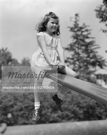 1950s 7 YEAR OLD GIRL WEARING DRESS SIT ON SEESAW TEETER TOTTER IN UP POSITION IN PARK SMILING FUN PLAYGROUND PLAY UP AND DOWN