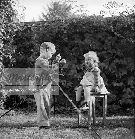 ANNÉES 1930 1940 BOY AVEC CAMÉRA SUR TRÉPIED PRISE PHOTO DE BÉBÉ FILLE PETIT ENFANT ASSIS SUR LE TABOURET