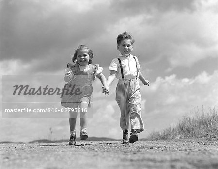 ANNÉES 50 ANNÉES 1940 GARÇON FILLE MAIN DANS LA MAIN QUI SILLONNE LE CIEL DE NUAGES DE CHEMIN DE TERRE DERRIÈRE HEUREUX SOURIANT COUPLE AMIS AMITIÉ AMOUR