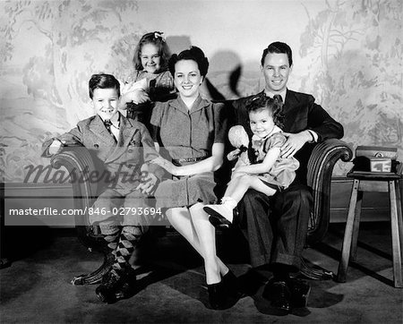 1940s FAMILY OF 5 PORTRAIT SITTING ON COUCH FACING CAMERA SMILING MOTHER FATHER TWO GIRLS ONE BOY WALLPAPER PRINT IN BACKGROUND