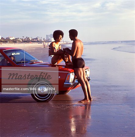 1970s SMILING ROMANTIC AFRICAN AMERICAN VACATION COUPLE AT BEACH WOMAN SITTING ON CAR HOOD MAN HOLDING HANDS