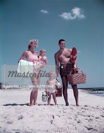 ANNÉES 1950 FAMILLE DEBOUT SANDY BEACH JOUETS PICNIC BASKET HOMME FEMME MÈRE PÈRE TROIS ENFANTS GARÇON FILLE