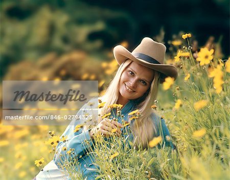 1970s RETRO BLOND WOMAN WEARING COWBOY HAT SITTING IN A FIELD OF BUTTERCUP WILD FLOWERS SMILING