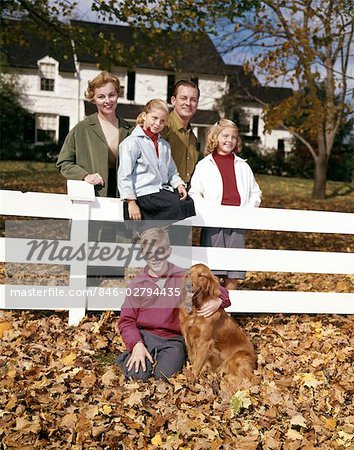 1960s PORTRAIT OF SMILING PARENTS WITH SON TWO DAUGHTERS AND DOG AT WHITE BOARD FENCE IN FRONT OF COLONIAL STYLE SUBURBAN HOUSE