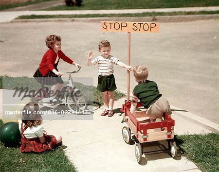 1950s BOYS AND GIRLS OUTDOOR WITH TRICYCLE AND WAGON PLAYING TRAFFIC WITH POLICE STOP SIGN AMERICANA