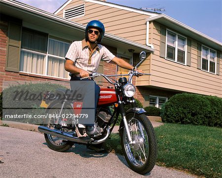 1970s YOUNG MAN BLUE HELMET SITTING ON MOTORCYCLE IN DRIVEWAY SUBURBAN HOUSE BIKE MOTORCYCLES MEN