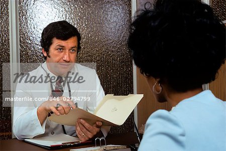 1970s AFRICAN AMERICAN WOMAN PATIENT CONSULTING WITH CAUCASIAN MAN DOCTOR IN OFFICE