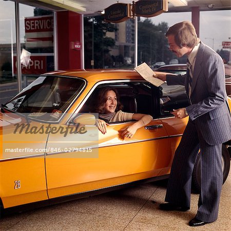 1970s SMILING WOMAN SITTING DRIVERS SEAT ORANGE CAR TALKING TO MAN AUTOMOBILE SALESMAN IN CAR DEALERSHIP