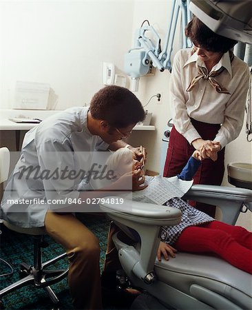 1970s MAN AFRICAN AMERICAN DENTIST WORKING ON LITTLE BLOND GIRL PATIENT WITH WOMAN MOTHER HOLDING HER HAND