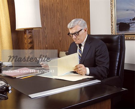 1960 1960s 1970 1970s MATURE MAN SILVER GRAY GREY HAIR EXECUTIVE SEATED DESK READING BUSINESS PAPERS MEN EXECUTIVES