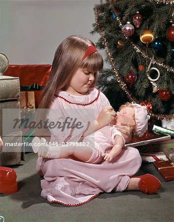 1970s YOUNG GIRL NIGHTGOWN AND SLIPPERS SITTING BY CHRISTMAS TREE GIVING DOLL BABY A BOTTLE