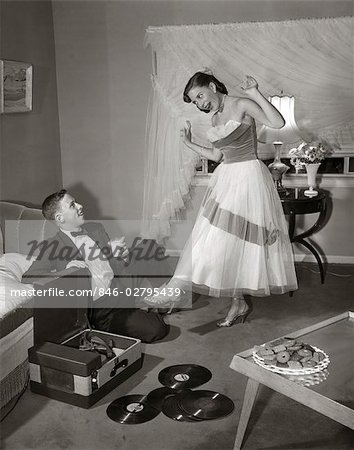 ANNÉES 1960 ANNÉES 1950 TEEN COUPLE IN LIVING ROOM IN PROM DRESS & SMOKING GUY ASSIS SUR LE PLANCHER & FILLE DANSANT SUR LES NOTES D'UN TOURNE-DISQUE